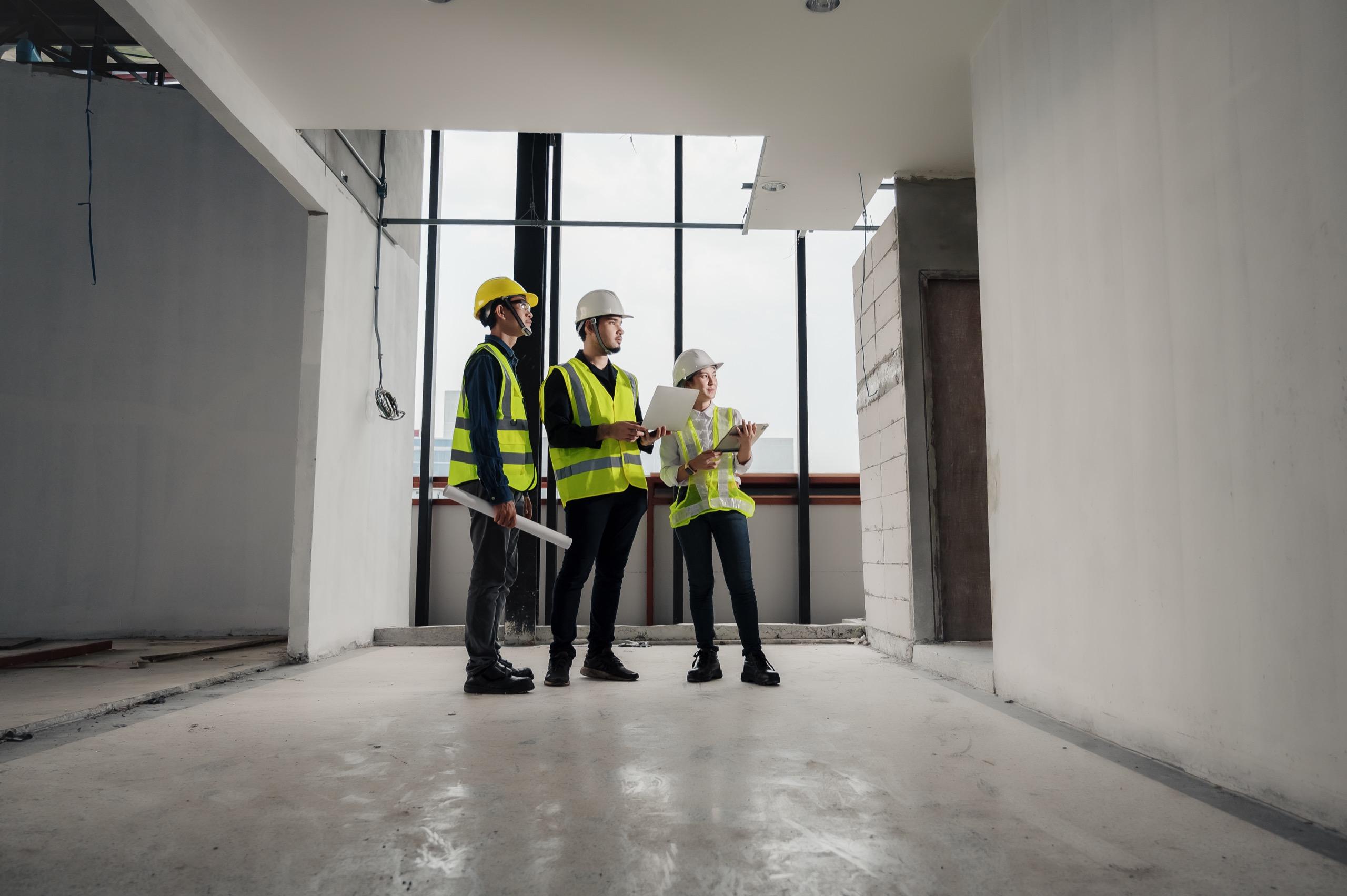 Group of construction professionals in hard hats and safety vests inspecting interior construction work with plans and a laptop.