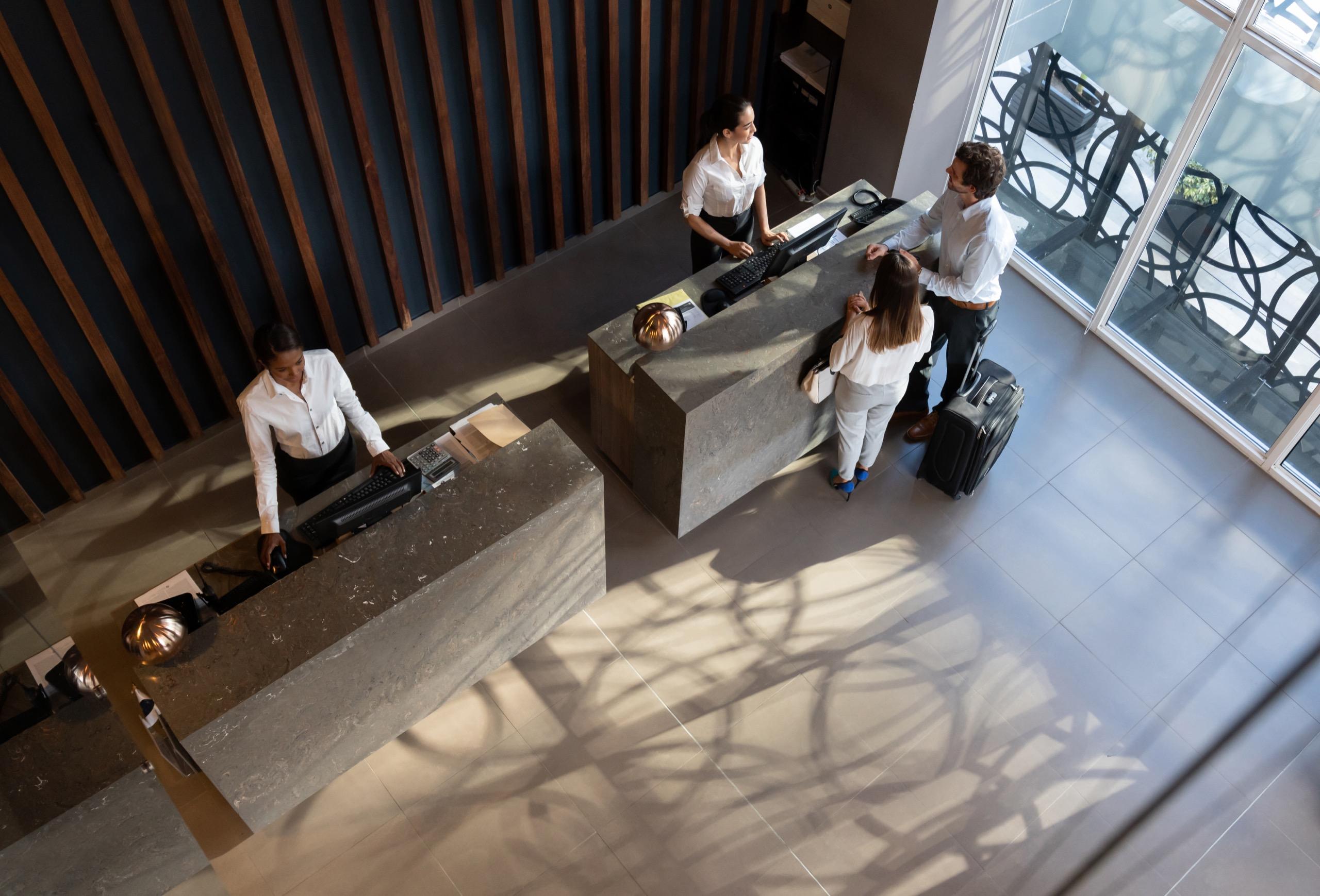 Receptionists assisting two guests with luggage at a modern front desk in a bright lobby.