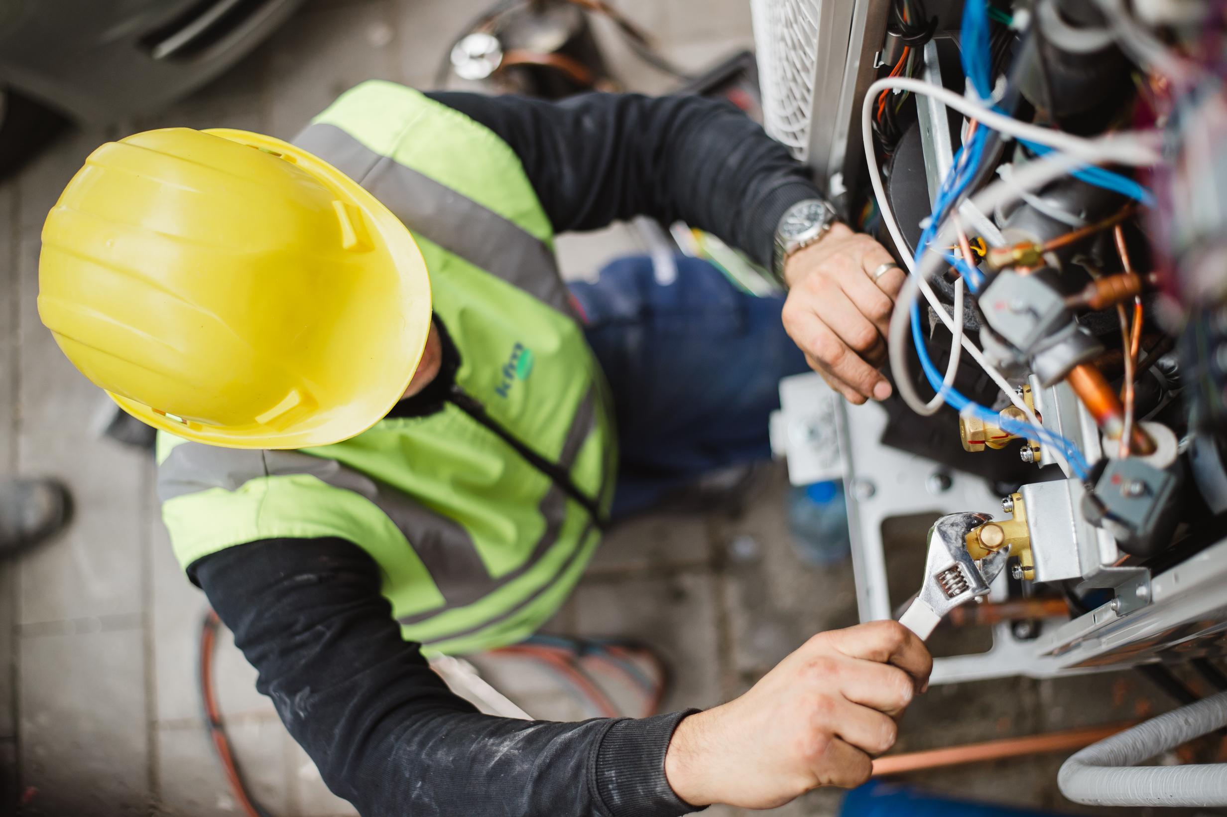 Technician wearing a yellow hard hat and high-visibility vest using a wrench to repair building mechanical components.