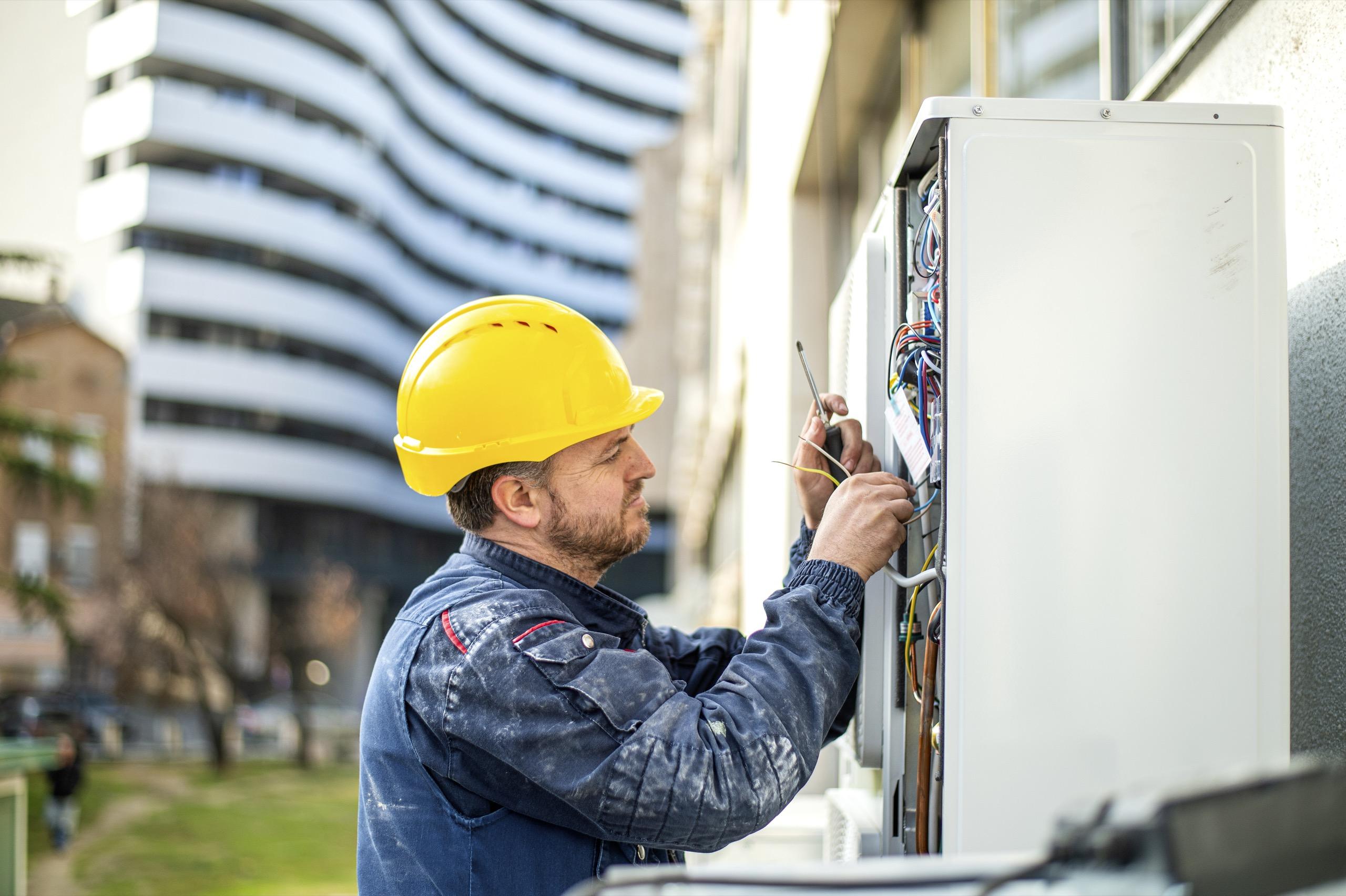 Technician wearing a yellow hard hat working on electrical wires inside an outdoor control panel.