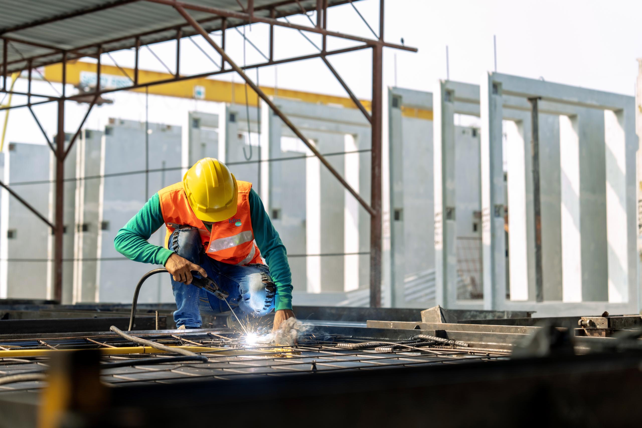 Worker in helmet and safety vest welding steel rods at a construction site.