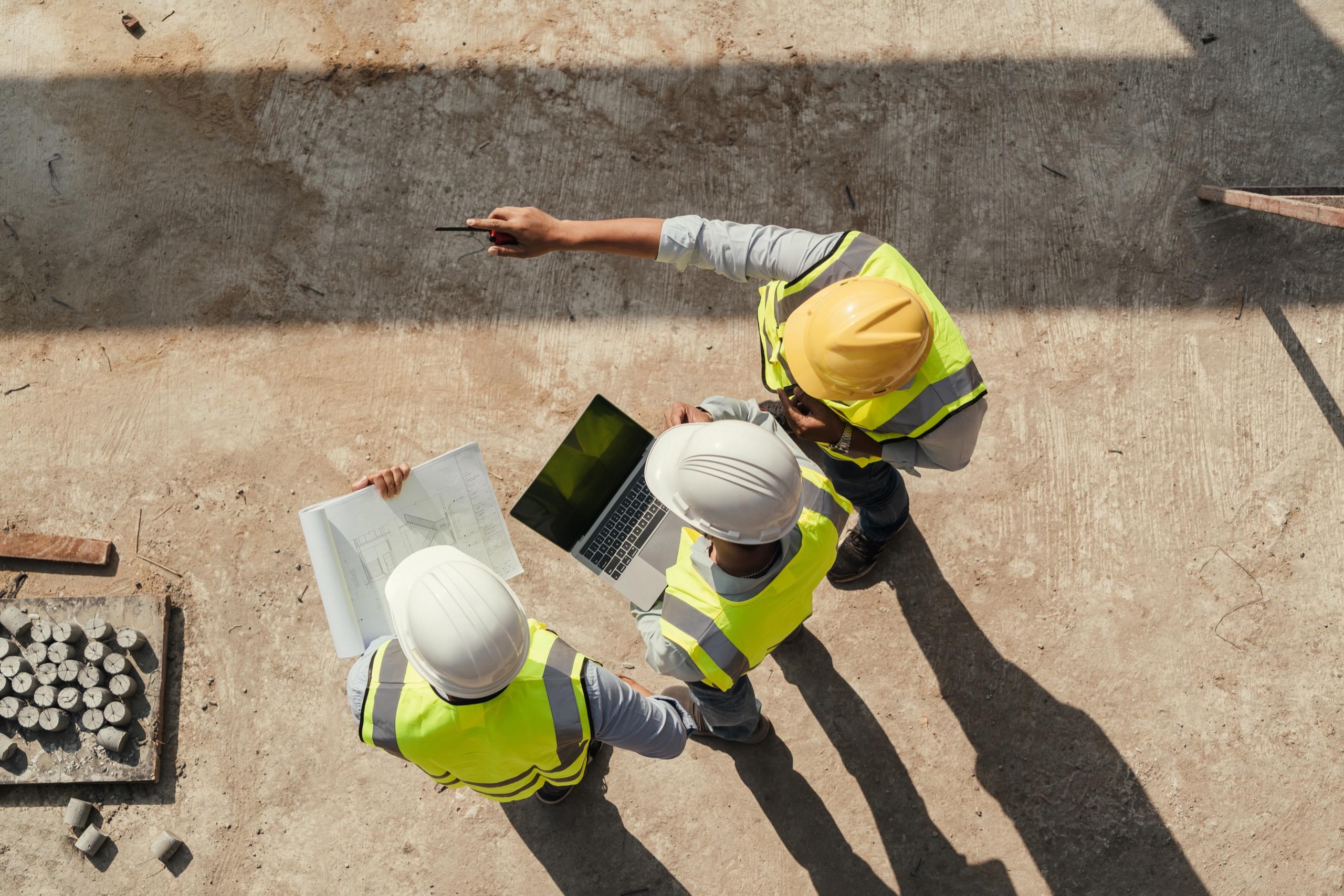 Construction workers in hard hats reviewing site plans with laptop and documents.