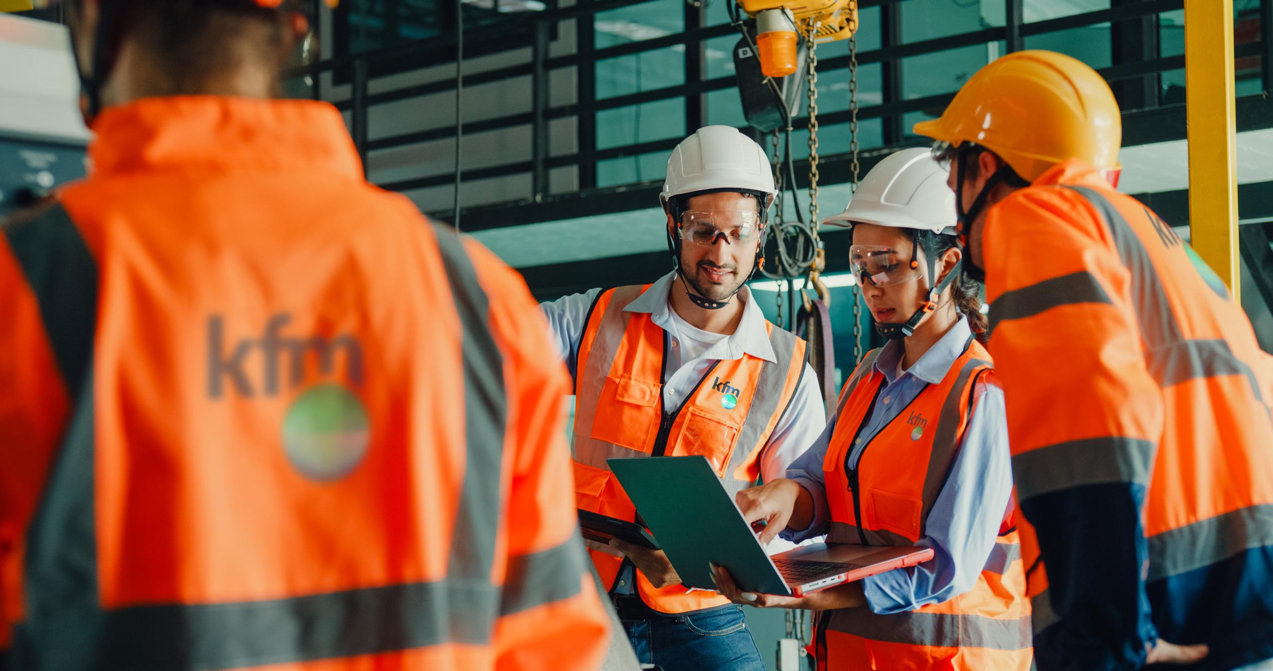 Engineers in hard hats and safety vests reviewing project plans on laptops at an industrial site.