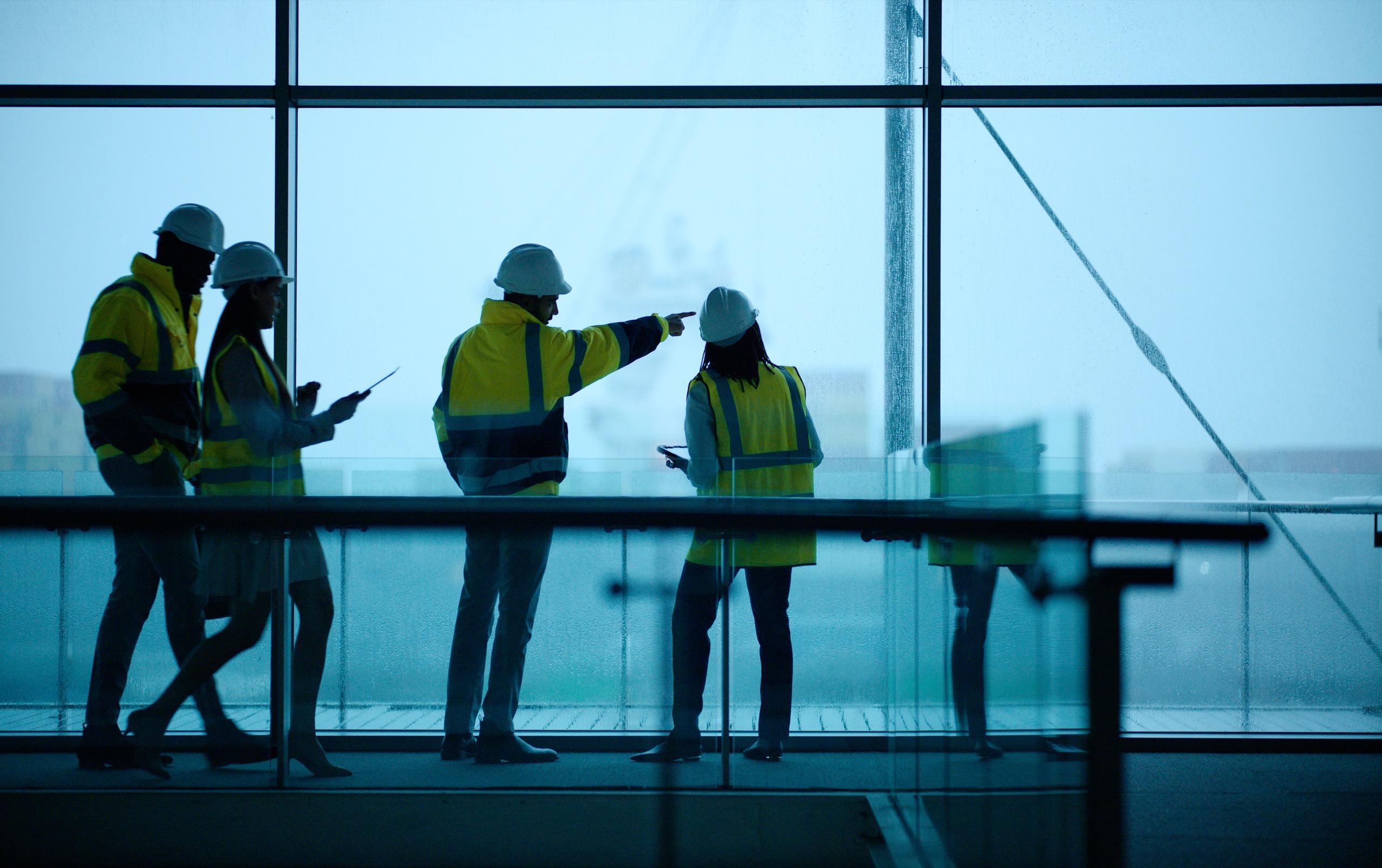 Silhouettes of construction team in hard hats and vests reviewing plans indoors.