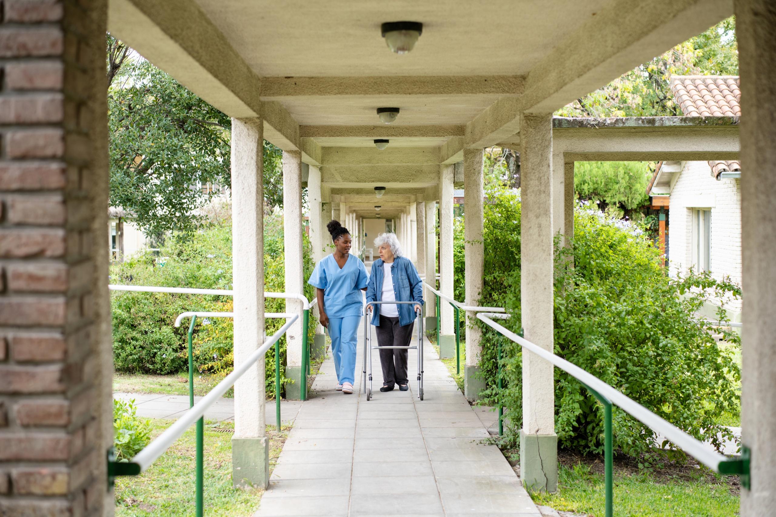 Nurse walking alongside an elderly woman with a walker in an outdoor aged care facility.