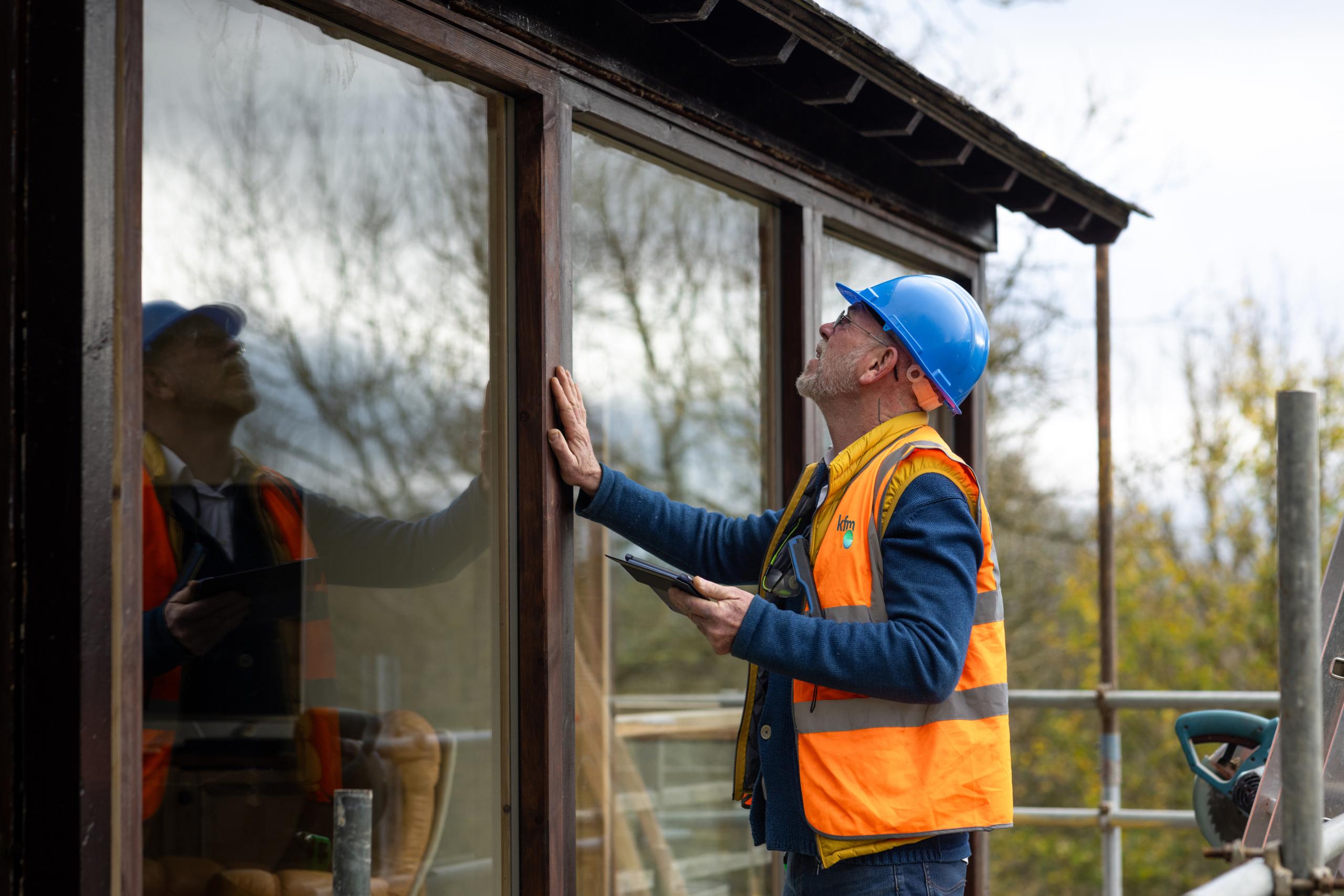 Construction inspector in hard hat checking wooden window frame with tablet in hand.
