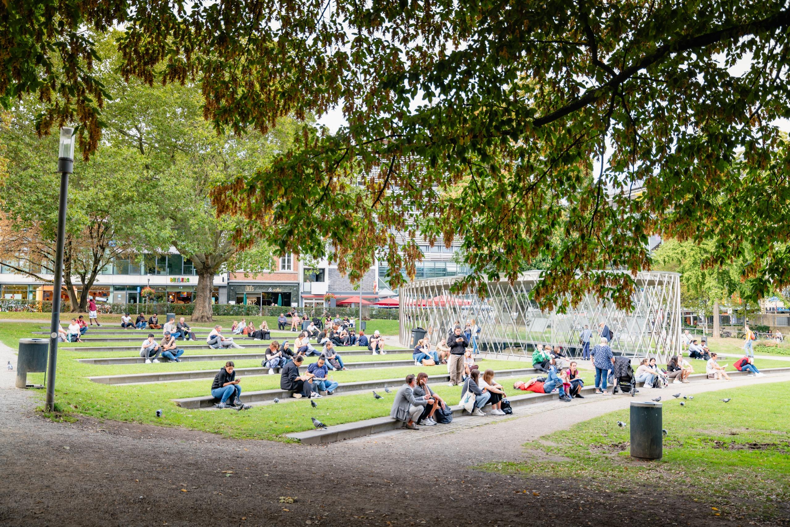 People sitting on steps and grass in a city park under large trees.