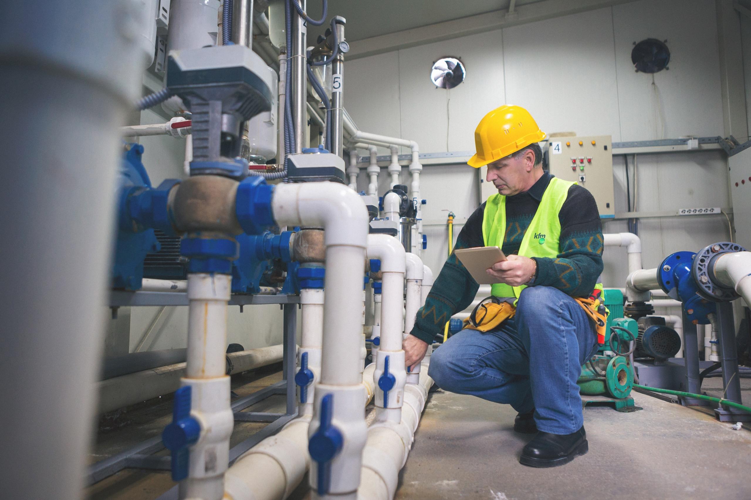 Engineer in helmet and vest checking water pipes in facility plant room.
